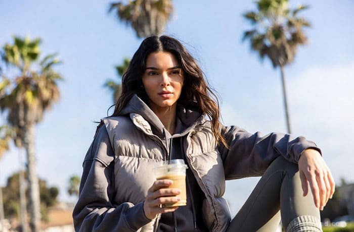 Lifestyle illustration of Kendall Jenner leaning against a car in Malibu, wearing a puffer jacket and looking effortlessly cool